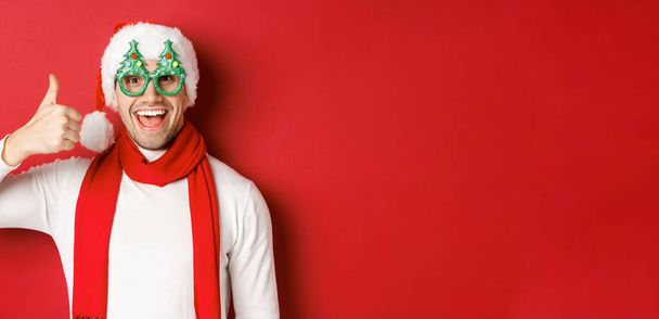 Koncepcja Bożego Narodzenia, ferie zimowe i uroczystości. Wesoły facet w czapce Mikołaja i okularach imprezowych, z kciukiem w górze i uśmiechem zadowolony, stojący na czerwonym tle - Zdjęcie, obraz