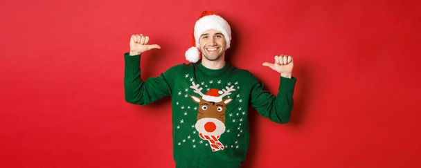 Begrip wintervakantie, kerst en levensstijl. Gelukkig aantrekkelijke man in santa hoed en trui, wijzend op zichzelf met een blij gezicht, staande over rode achtergrond - Foto, afbeelding