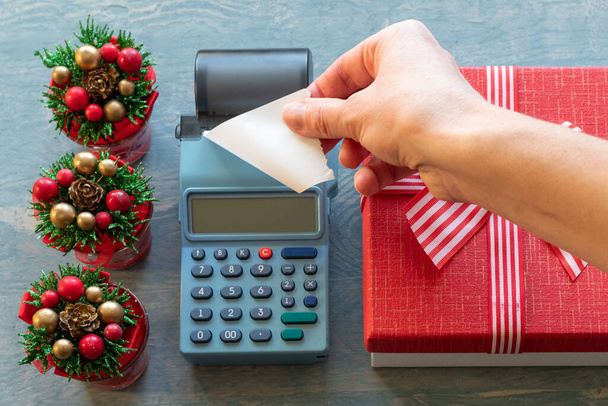 Christmas sale. Hand holding A blank white check from a cash register near a red gift box with a ribbon and small Christmas trees with cones. Buying gifts for New Year and Christmas - Foto, immagini