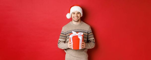 Πορτρέτο του όμορφου άνδρα που κρατά ένα δώρο, ευχόμενος καλές διακοπές, στέκεται σε καπέλο Σάντα και χριστουγεννιάτικο πουλόβερ σε κόκκινο φόντο - Φωτογραφία, εικόνα