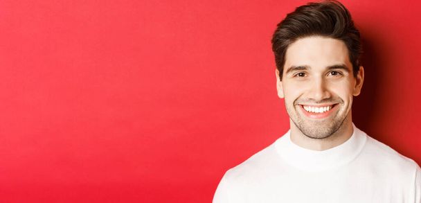 Großaufnahme eines gutaussehenden, lächelnden brünetten Mannes, der einen weißen Pullover trägt, glücklich und selbstbewusst lächelt und vor rotem Hintergrund steht - Foto, Bild