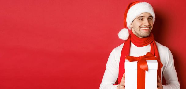 Concetto di vacanze invernali, Natale e stile di vita. Primo piano di uomo attraente in cappello e sciarpa di Babbo Natale, tenendo regalo di Capodanno, guardando a destra e sorridente, in piedi su sfondo rosso - Foto, immagini