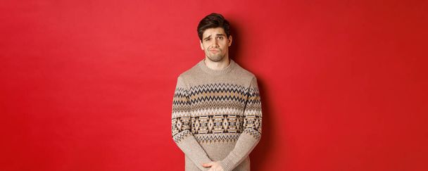 Portrait of gloomy and sad caucasian man in christmas sweater, frowning and pouting indecisive, having bad new year, standing over red background - Photo, Image