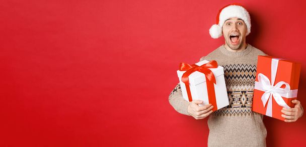 Εικόνα του χαρούμενου ελκυστικού άντρα κρατώντας χριστουγεννιάτικα δώρα, στέκεται στο καπέλο Σάντα και το χειμερινό πουλόβερ, χαμογελώντας κατάπληκτος, στέκεται πάνω από το κόκκινο φόντο - Φωτογραφία, εικόνα