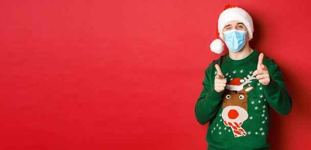 Concepto de año nuevo, covidio-19 y distanciamiento social. Hombre alegre en suéter de Navidad, máscara médica y sombrero de santa, señalando los dedos a la cámara, deseando felices fiestas, de pie sobre el fondo rojo - Foto, Imagen