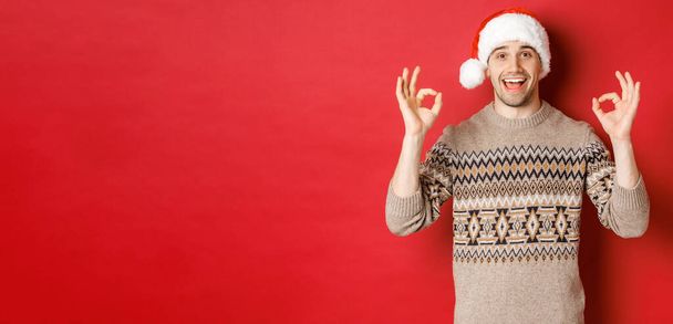 Portret szczęśliwego i zadowolonego przystojniaka, ubrany w zimowy sweter i czapkę Mikołaja, z dobrymi znakami, chwała dobrej imprezie świątecznej, stojący nad czerwonym tłem - Zdjęcie, obraz