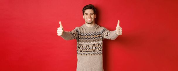 Beeld van knappe blanke man in kersttrui, met duimen omhoog in goedkeuring en glimlach, wens fijne feestdagen, staande over rode achtergrond - Foto, afbeelding