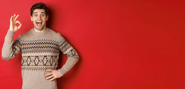 Concept of christmas celebration, winter holidays and lifestyle. Amazed and happy man in xmas sweater, showing okay sign and looking at something fantastic, red background - Foto, Imagem
