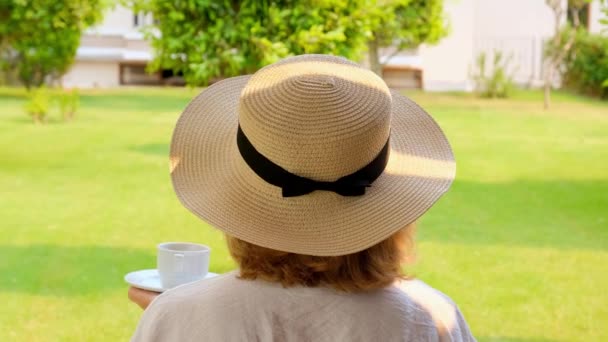 Senora femmina, 50-55 anni in un cappello di paglia tiene una tazza di bevanda calda e beve caffè, seduta al mattino su una terrazza estiva in una giornata di sole. Il concetto di buongiorno, viaggio e aspettativa di - Filmati, video