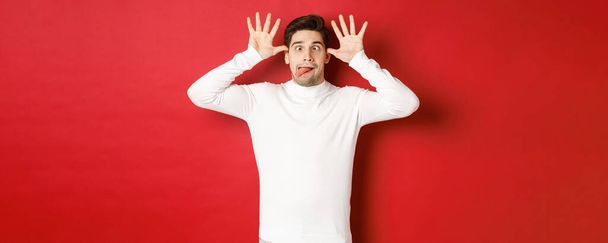 Retrato de tipo caucásico divertido, mostrando la lengua y haciendo caras, usando suéter blanco, de pie sobre fondo rojo - Foto, imagen