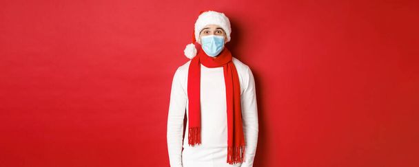 Concept de nouvel an, coronavirus et vacances. Homme joyeux célébrant Noël et distanciation sociale, portant un masque médical, un chapeau et une écharpe de Père Noël, debout sur fond rouge - Photo, image