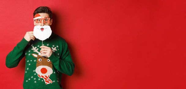 Πορτρέτο του έκπληκτος άνθρωπος σε πράσινο χριστουγεννιάτικο πουλόβερ, κρατώντας αστεία μάσκα Santa Claus, γιορτάζει το νέο έτος, στέκεται πάνω από το κόκκινο φόντο - Φωτογραφία, εικόνα