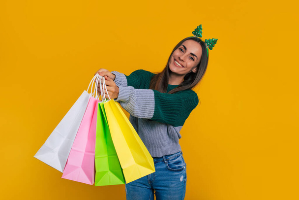 Funny attractive young brunette woman in Christmas shopping time with colorful paper bags for purchase and gifts isolated on yellow background - Valokuva, kuva