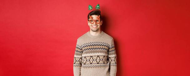 Portrét pohledného vousatého chlapa ve slavnostních brýlích a zimním svetru, s potěšením se usmívá, slaví nový rok, přeje veselé Vánoce, stojí nad červeným pozadím - Fotografie, Obrázek