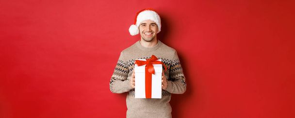Εικόνα ενός όμορφου χαμογελαστού άντρα με καπέλο και χειμωνιάτικο πουλόβερ, που κρατάει ένα δώρο, δίνει χριστουγεννιάτικο δώρο και εύχεται καλές γιορτές, στέκεται πάνω από το κόκκινο φόντο - Φωτογραφία, εικόνα