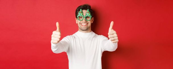 Zbliżenie atrakcyjnego szczęśliwego mężczyzny w okularach imprezowych i białym swetrze, z kciukami do góry w aprobacie i uśmiechem, stojącego na czerwonym tle - Zdjęcie, obraz