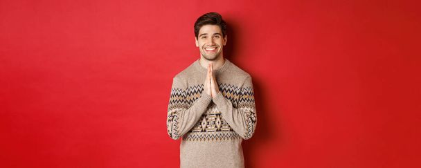 Imagem de homem feliz e bonito em camisola de Natal, pedindo por favor, de mãos dadas, sorrindo e dizendo obrigado, sentindo-se grato, de pé sobre fundo vermelho - Foto, Imagem