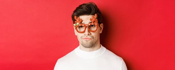 Έννοια των χειμερινών διακοπών, τα Χριστούγεννα και γιορτή. Κοντινό πλάνο του δύσπιστου όμορφου άντρα, που φοράει γυαλιά για πάρτι, χαμογελάει και δείχνει ασυγκράτητος, στέκεται πάνω από το κόκκινο φόντο - Φωτογραφία, εικόνα