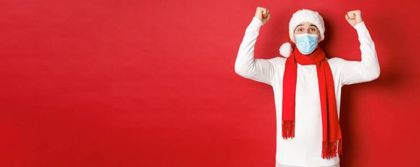Concept of covid-19, christmas and holidays during pandemic. Portrait of happy man in santa hat and medical mask, rejoicing and celebrating new year, standing over red background - 写真・画像