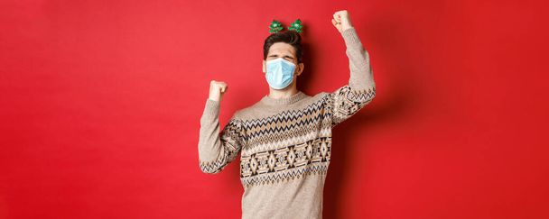 Concept van coronavirus, wintervakantie en sociale afstand. Beeld van opgewonden en gelukkige man in medisch masker en kerstkleding, handen omhoog steken en nieuwjaar vieren, rode achtergrond - Foto, afbeelding