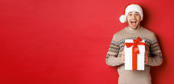 Bild eines aufgeregten gutaussehenden Mannes, der Weihnachtsgeschenk erhält, Weihnachtsmütze und Winterpullover trägt, vor Freude schreit, Geschenk hält und über rotem Hintergrund steht - Foto, Bild