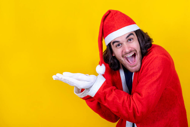 Un giovane sorridente vestito da Babbo Natale indica il tuo messaggio con le mani. Sfondo giallo e spazio di copia per il messaggio di saluto. - Foto, immagini