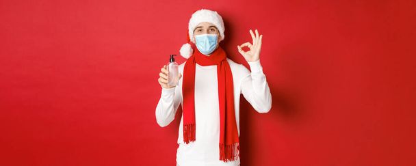 Concept of covid-19, christmas and holidays during pandemic. Attractive man in santa hat and medical mask, showing okay sign while recommending hand sanitizer, standing over red background - Photo, Image