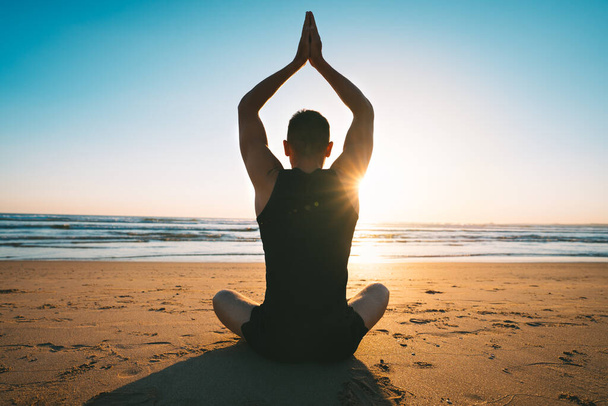 Man sitting on the sand and practise yoga or meditates on the beach with ocean view at sunset or sunrise - Photo, Image