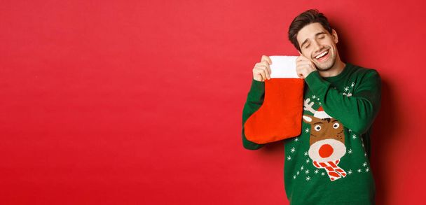 Portrait of happy attractive man in green sweater, looking delighted to receive gifts in christmas stocking, standing over red background - Photo, Image