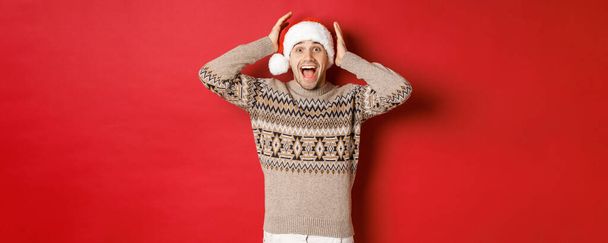Bild eines überraschten und glücklichen jungen Mannes mit Weihnachtsmütze und Weihnachtspullover, der ein tolles Geschenk erhält und aufgeregt vor rotem Hintergrund steht - Foto, Bild