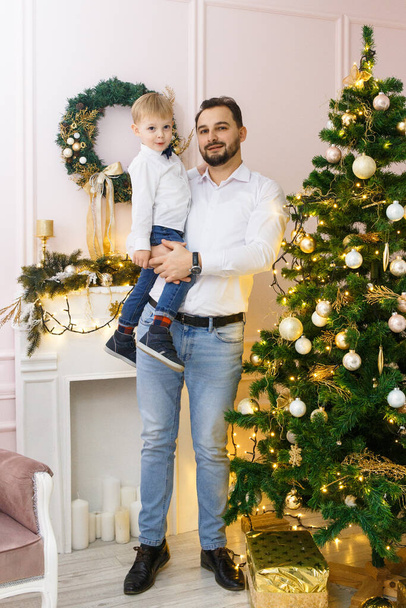Happy Father and son against Christmas tree with Christmas balls and light garland preparing for celebrating winter holidays: Merry Christmas Eve and Happy New Year 2021. Family concept. - Фото, изображение