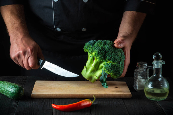 Professional chef cuts fresh Broccoli with a knife for salad on vintage kitchen table with fresh vegetables. Cooking and restaurant or cafe concept - Foto, Imagem