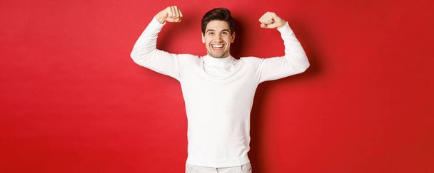Πορτρέτο του χαμογελαστού όμορφος άντρας σε λευκό πουλόβερ, κάμψη δικέφαλους και κομπορρημοσύνη με δύναμη, επίδειξη ισχυρή μυς μετά την προπόνηση, στέκεται πάνω από το κόκκινο φόντο - Φωτογραφία, εικόνα