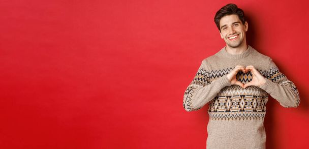 Image of handsome bearded guy in winter sweater, wishing merry christmas and showing heart sign, smiling lovely at camera, standing against red background - Photo, Image