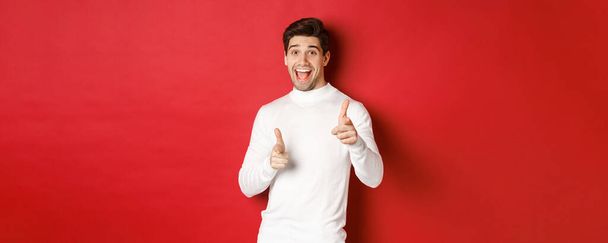 Concept of winter holidays, christmas and lifestyle. Cheerful handsome guy in white sweater congratulating you, pointing fingers at camera and wishing happy new year, standing over red background - Photo, Image