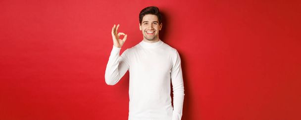 Pojęcie ferii zimowych, Bożego Narodzenia i stylu życia. Szczęśliwy przystojny mężczyzna w białym swetrze z dobrym znakiem, uśmiechnięty zadowolony, polecam sklep, stojący na czerwonym tle - Zdjęcie, obraz