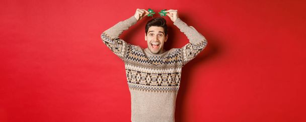 Concept of winter holidays, christmas and celebration. Image of funny and cute adult man enjoying new year party, smiling and rejoicing, standing over red background - Zdjęcie, obraz