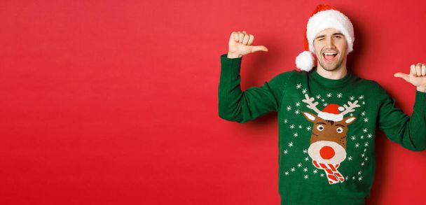 Concepto de vacaciones de invierno, Navidad y estilo de vida. Hombre guapo descarado en sombrero de santa y suéter verde, apuntándose a sí mismo y guiñando el ojo, de pie sobre el fondo rojo - Foto, imagen