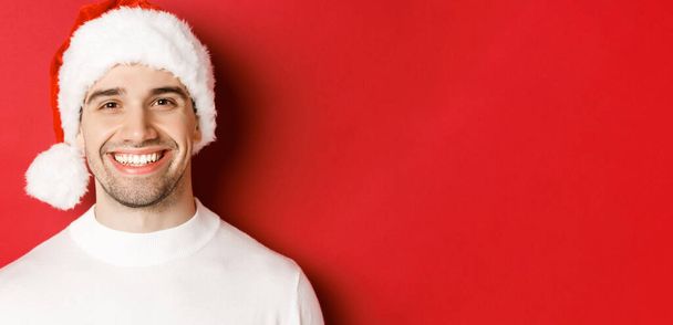 Primer plano del atractivo hombre sonriente en jersey blanco y sombrero de santa, luciendo feliz, disfrutando de las vacaciones de invierno, de pie sobre fondo rojo - Foto, imagen