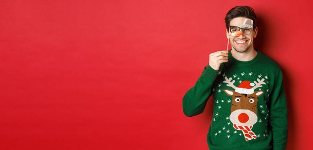 Homem engraçado em camisola de Natal e máscara de festa, celebrando férias de inverno, sorrindo feliz, de pé sobre fundo vermelho - Foto, Imagem