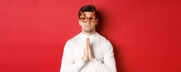 Крупный план симпатичного парня в рождественских очках, умоляющего о помощи, нуждающегося в услуге, стоящего на красном фоне - Фото, изображение