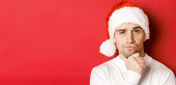 Close-up of thoughtful handsome man in santa hat, frowning and looking at camera, thinking about something, standing over red background - Foto, Imagen