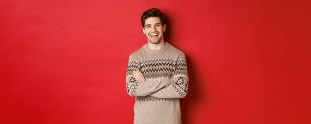 Retrato de homem alegre e atraente em camisola de Natal, rindo e sorrindo, celebrando feriados de ano novo e inverno, em pé sobre fundo vermelho - Foto, Imagem