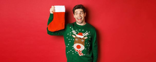 Image d'un homme surpris et amusé en pull vert, regardant les bas de Noël avec des cadeaux et souriant, debout sur fond rouge - Photo, image