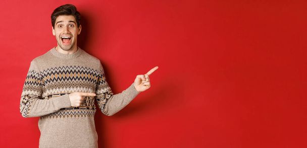 Concept of christmas celebration, winter holidays and lifestyle. Handsome man in xmas sweater pointing fingers right, smiling amazed, showing new year promo against red background - Fotoğraf, Görsel
