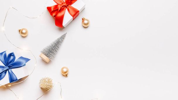 Decorazione natalizia. Regalo bianco con fiocco rosso, palline d'oro e ghirlanda di luci scintillanti nella decorazione natalizia su sfondo bianco per biglietto di auguri. Layout piatto, vista dall'alto, spazio di copia - Foto, immagini