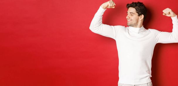 Портрет улыбающегося красавца в белом свитере, сгибающего бицепсы и хвастающегося силой, демонстрирующего сильные мышцы после тренировки, стоящего на красном фоне - Фото, изображение