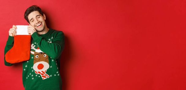 Portrait of happy attractive man in green sweater, looking delighted to receive gifts in christmas stocking, standing over red background - Photo, Image