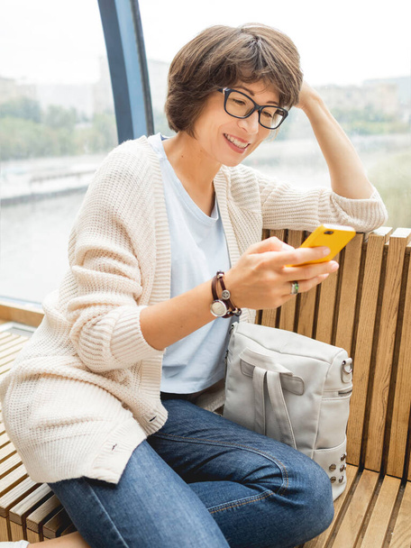 Smiling woman in eyeglasses texting on smartphone. Millennials lifestyle. Casual clothes. - Fotó, kép