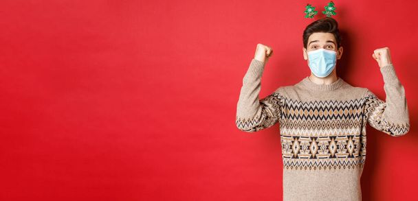Conceito de Natal, feriados e coronavírus. Homem feliz celebrando o ano novo durante o surto de covid-19, usando máscara médica e suéter, regozijando-se com o fundo vermelho - Foto, Imagem